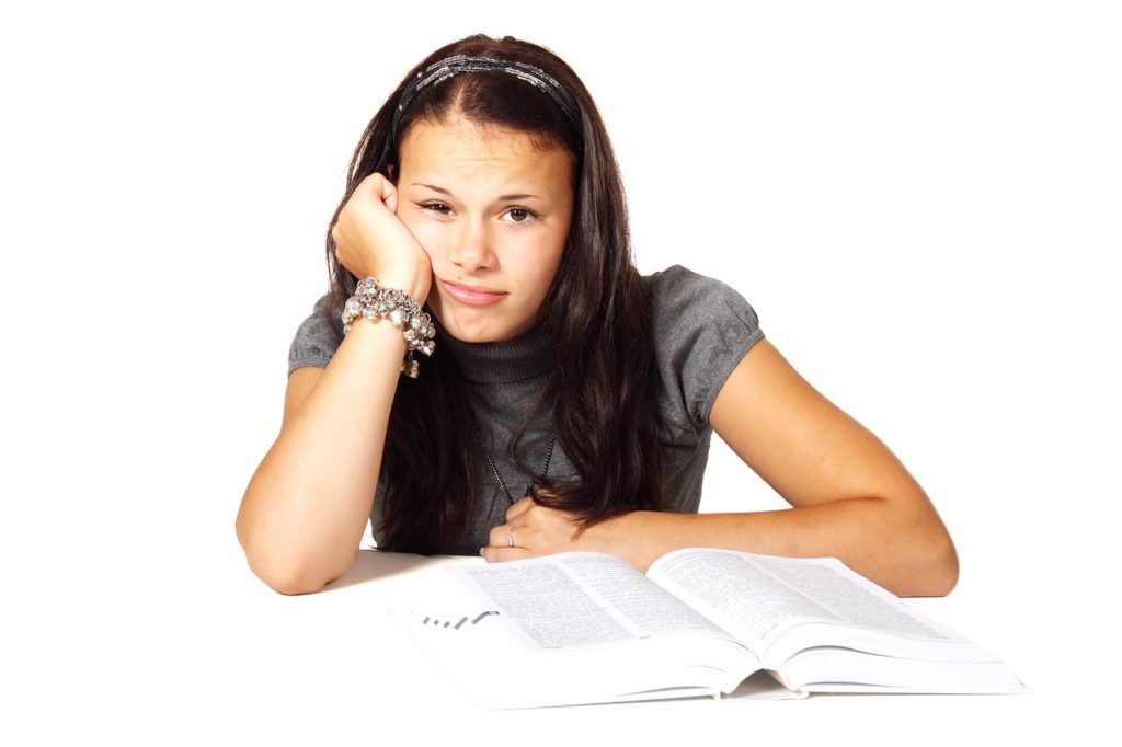 Young write girl with an open book in front of her, right palm on right cheek, bored face.